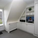 A room with sloped ceilings, window, TV, and white cabinets.