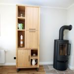Hölzerner Schrank mit offenen Regalen und Türen. Schwarzer Kaminofen daneben. Tisch und Stuhl links.