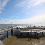 Harbor promenade with pier and flags over water. People walk along the waterfront.