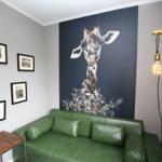 Ein Raum mit grünem Sofa, Wandbild einer Giraffe und einer Lampe in Form eines Straußens.