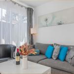 Ein Wohnzimmer mit grauem Sofa, blauen Kissen und einem schwarzen Sessel. An der Wand hängt ein Bild.