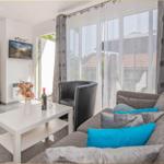 Wohnzimmer mit Sofa, Couchtisch und TV. Große Fenster mit Blick auf Außenbereich.