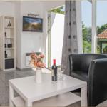 Wohnbereich mit schwarzer Couch, weißem Tisch und TV. Durchgehende Glasfront mit Blick auf Garten.