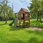 Großer Garten mit Holzspielhaus, Rutsche und Schaukel unter Bäumen.