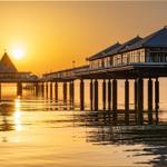 Sonnenuntergang über einem Steg mit Pavillon und Reflexion im Wasser.