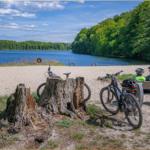 Zwei Radfahrer sitzen auf einer Bank am Strand eines Sees mit Bäumen im Hintergrund.