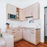 Kitchen with wooden cabinets and microwave. Living area with sofa. Wooden floor and white walls.