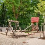 Kinderspielplatz mit Schaukel, Rutsche und Klettergerüst. Umgeben von Bäumen und gepflastertem Weg.