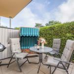 Terrace with table, chairs, grill, and lounge chair behind hedge.