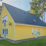 Ein gelbes Haus mit dunklem Dach, umgeben von Gras und Bäumen. Die Tür und Fenster sind weiß.