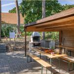 Holzüberdachter Bereich mit Grill, Tisch und Bänken. Im Hintergrund Spielplatz und Baum.
