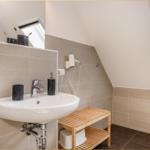 Badezimmer mit Waschbecken, Spiegel, Handtuchhalter und Holzbank unter Dachschrägen.