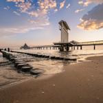Strand mit Steinen, Pier und Leuchtturm bei Sonnenuntergang.