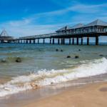 Strand mit Wellen, Steine im Wasser und Pier mit Dächern und Besuchern.