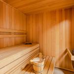 Sauna mit Holzbank, Eimer und Holzhammer, Wanduhr und Kachelofen sichtbar.
