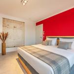 Schlafzimmer mit roter Wand, Bett mit Kissen und Decke, Wandbild und Stehlampe.