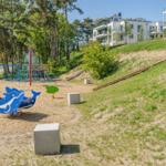 Kinderspielplatz mit Delfin-Schaukel und Klettergerüst. Hintergrund: Wohngebäude auf grüner Hanglage.