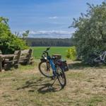 Zwei E-Bikes stehen auf einer Wiese neben einem Holztisch und Bänken. Im Hintergrund sind Felder und Bäume.