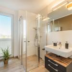 Badezimmer mit Dusche, Waschbecken und Fenster. Holzboden und Wandfliesen.