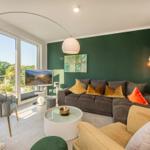 Wohnzimmer mit grüner Wand, Sofa, Sessel und TV. Große Fenster mit Blick auf Garten.