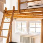 Upper bunk bed with wooden frame and ladder. Two windows and radiator on the wall.