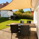Terrasse mit Tisch und Stühlen unter Sonnenschutz, umgeben von Grünfläche und Hecke.