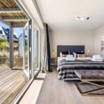 Schlafzimmer mit Blick auf Terrasse und Grünflächen. Bett mit Kissen und Decke. Wandbild und Sitzbank.