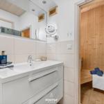 Bad mit Waschbecken, Spiegel und angrenzender Sauna mit Holzwand.