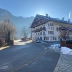 Haus mit Balkonen und Schornsteinrauch. Schnee liegt am Straßenrand. Berg im Hintergrund.