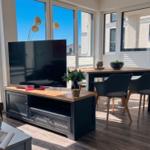 Wohn- und Essbereich mit TV, Tisch und Stühlen. Große Fenster mit Blick auf Gebäude.
