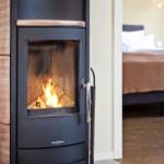 Kaminofen mit brennendem Holz, daneben Schlafzimmer mit Bett und Holzboden.