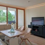 Wohnzimmer mit Sofa, Esstisch, Fernseher und großem Fenster mit Blick auf Bäume.