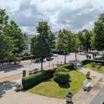 Aussicht auf grünen Platz mit Bäumen, Beeten und Gehweg. Menschen gehen und Autos parken.