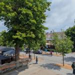 Straßenaussicht mit Bäumen, Café und Passanten. Steinpflaster und Parkplatz sind sichtbar.