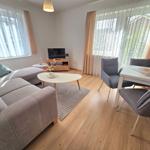 Wohnzimmer mit Sofa, Esstisch und Fernseher. Holzboden und große Fenster.