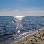 Strand mit sanften Wellen und Sonnenreflexionen auf dem Wasser unter blauem Himmel.