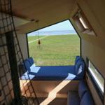 Blick aus Holzhaus auf Grasland und Meer mit blauem Fensterbanksofa.