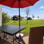 Terrace with table and chairs under a red umbrella. View of green grass and houses by the water.