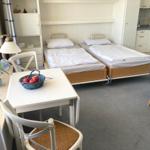 Two beds, table with chairs, kitchen area, and window with curtains.