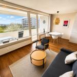 Living area with sofa, table, and chair. Large windows with view of balcony and city.