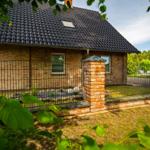 Haus mit Ziegelfassade und Dachgaube hinter Zaun mit Steinpfeilern.