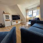 Wohnzimmer mit blauem Sofa, TV und Holzboden. Fenster mit Vorhängen.