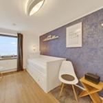 Schlafzimmer mit weißem Bett, lila Wand und Holzboden. Fenster mit Blick auf Wasser.