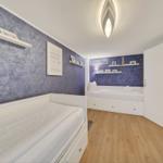 Zwei weiße Betten mit Schubladen, lila Wand, Holzboden und Deckenleuchte.