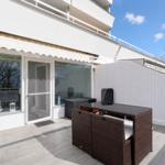 Terrace with seating area, glass front, and white wall. Sky is blue with white clouds.