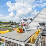 Terrasse mit Tisch, Stühlen und Grill. Blick auf Ferienhäuser und Grünflächen.