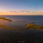 Aerial view of coastal area with lighthouse, marina, and green dunes at sunset.