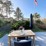 Holztisch mit Stühlen auf Terrasse, Blumen, Flagge und Bäume im Hintergrund.