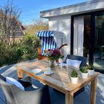 Terrasse mit Holztisch, Stühlen und Strandkorb. Blumen und Pflanzen schmücken den Außenbereich.
