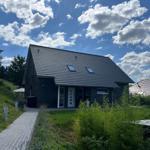 Ein modernes Haus mit dunklem Dach und weißen Fensterrahmen. Ein gepflasterter Weg führt zum Eingang.